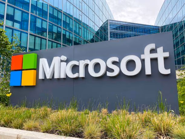 Microsoft uzņēmuma logo un nosaukums uz pelēkas plāksnes modernas stikla biroju ēkas priekšplānā.