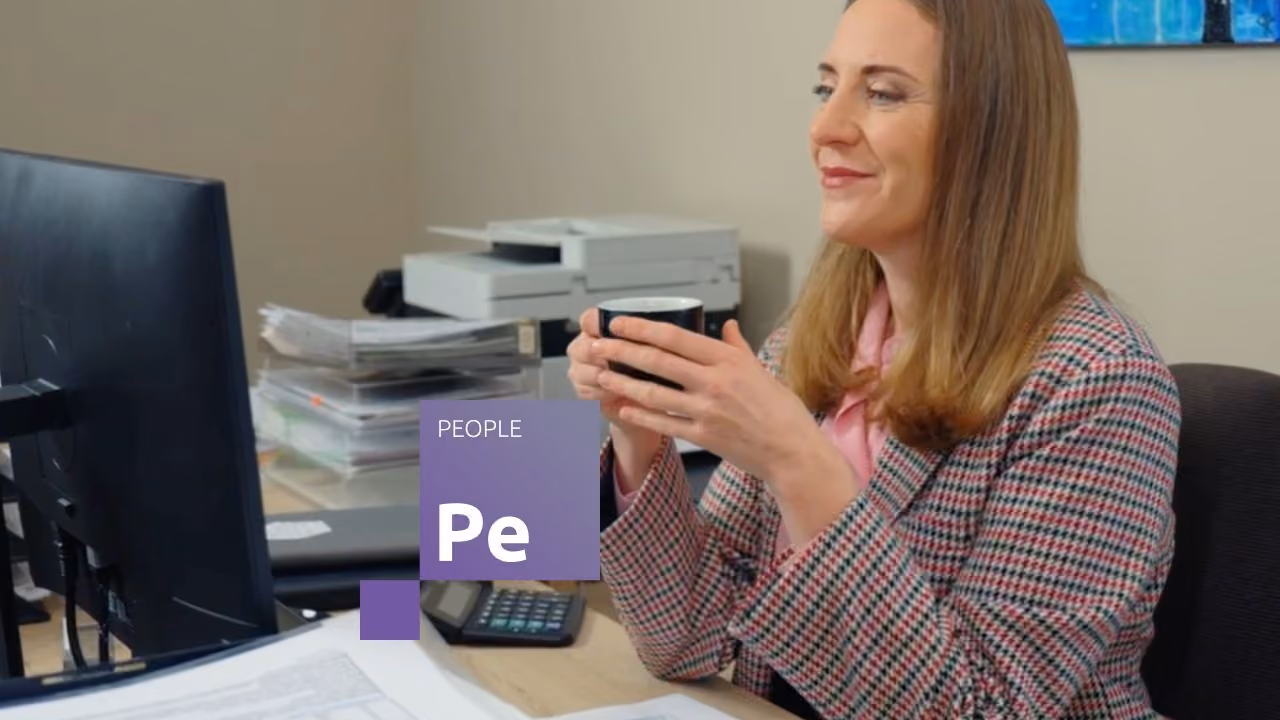 Smaidīga darbiniece birojā, turot kafijas krūzi un izmantojot Jumis People risinājumu darbam pie datora.