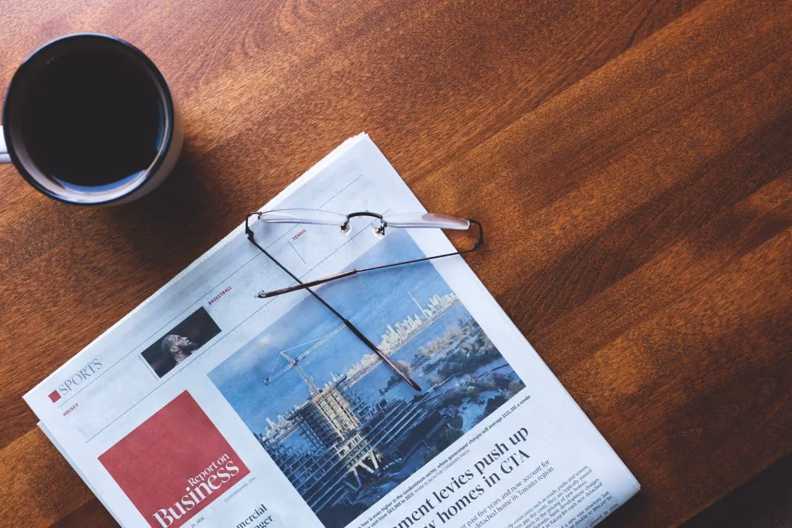 Skats no augšas uz koka galda esošu avīzi "Report on Business", brillēm un tasi melnas kafijas.
