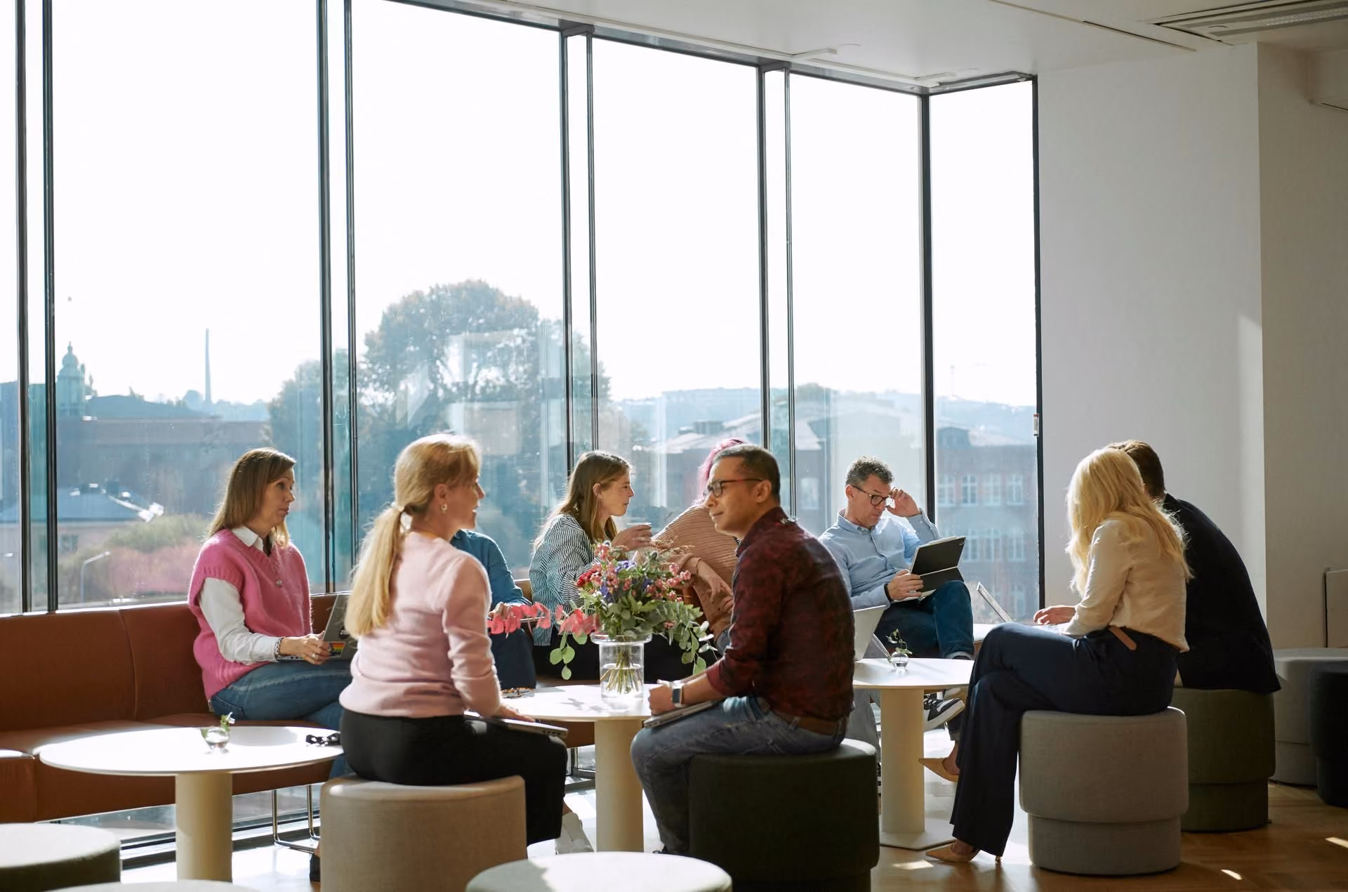 A group of people talking and working at a table in a bright, modern office with large windows.