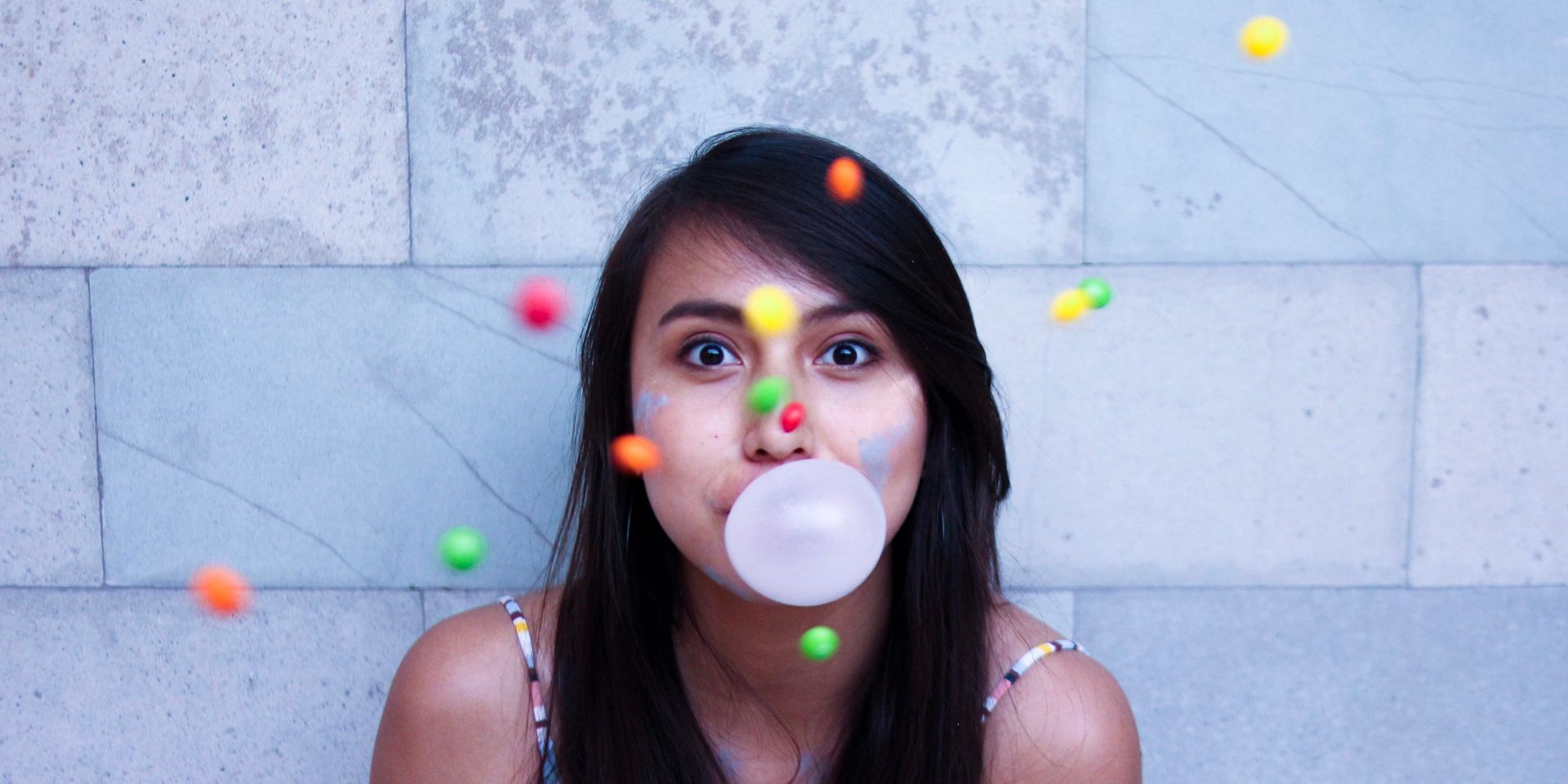 A person blows bubblegum while colourful sweets fly through the air against a light stone wall.