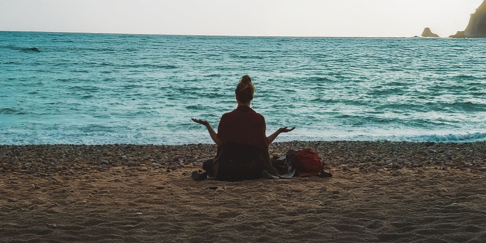 A person sits on a beach with their back to the camera, looking out at the sea and sunset.