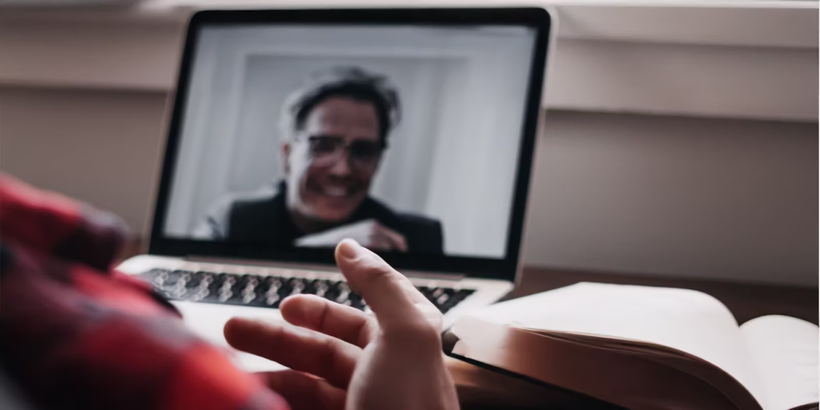 Persona piedalās videozvanā, sarunājoties ar vīrieti klēpjdatora ekrānā; blakus datoram atrodas atvērta piezīmju grāmata.