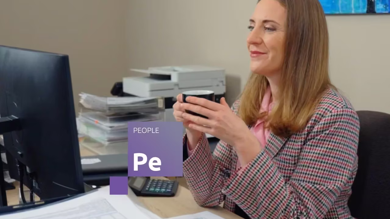 Smaidīga darbiniece birojā, turot kafijas krūzi un izmantojot Jumis People risinājumu darbam pie datora.