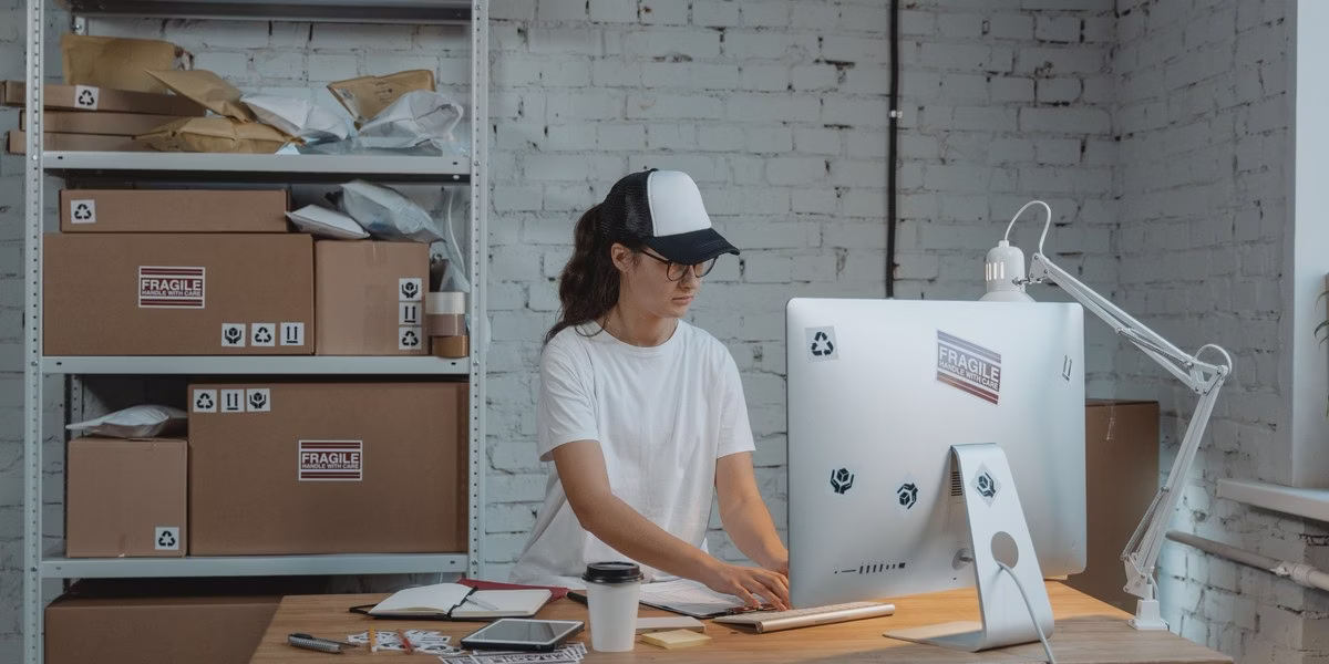 An e-commerce business employee processes orders on a computer while working in a warehouse surrounded by shipping packages.
