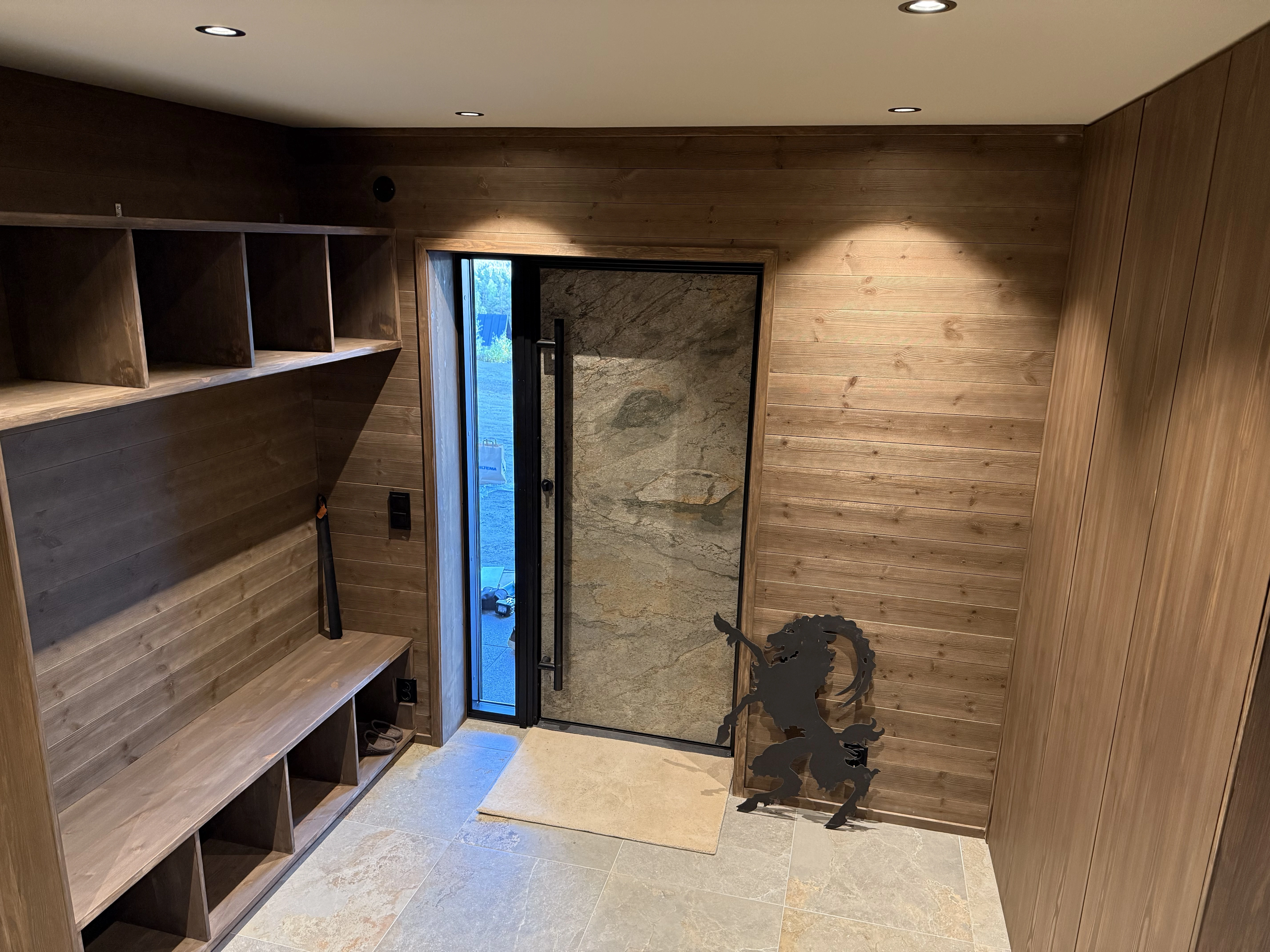 Träpanel-entrén med inbyggda trämöbler och bänk, stengolv, stendörr med svart ram .