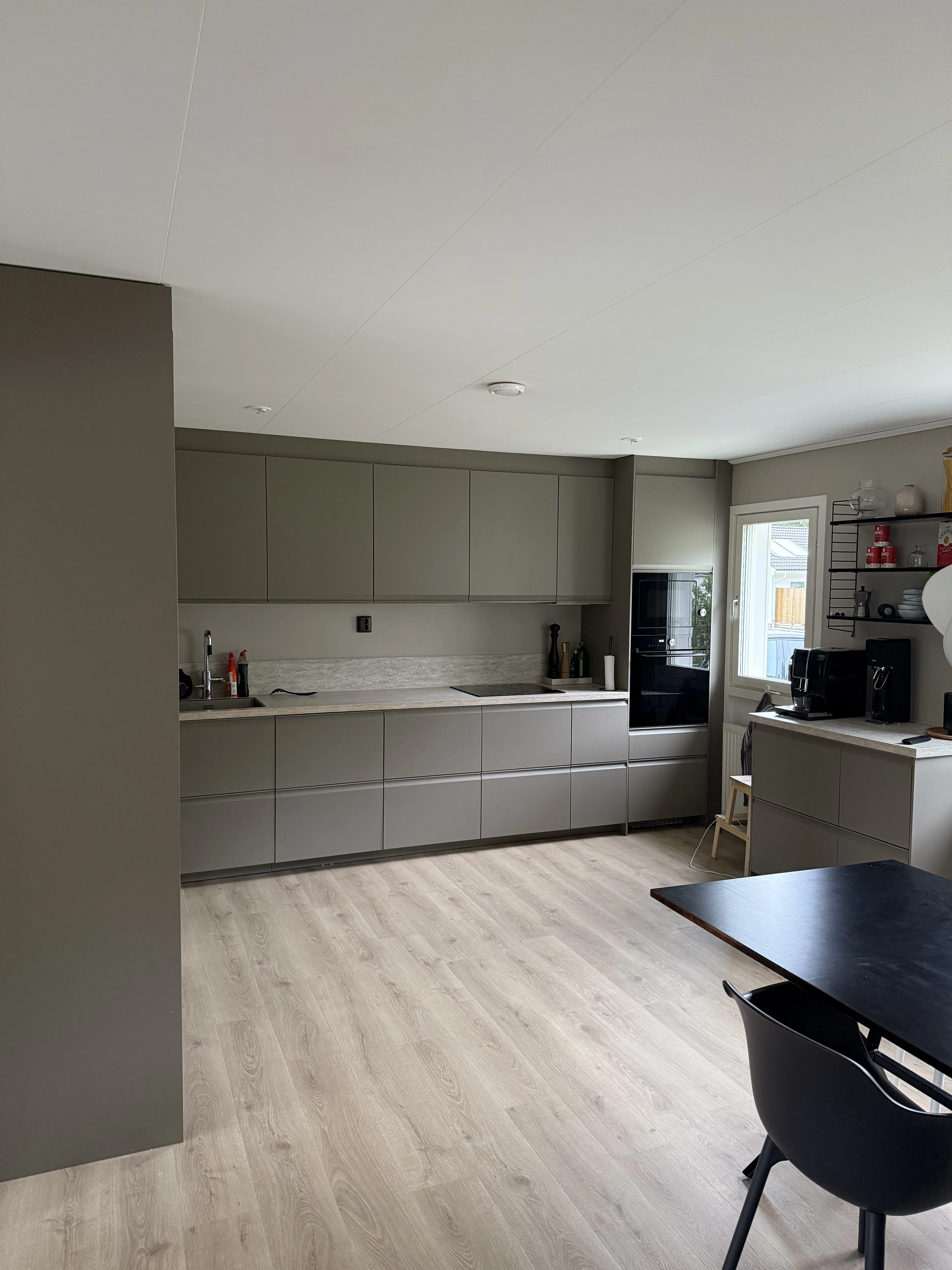 Modernt kök med mattgrå skåpsluckor, ljust trägolv, svart matbord och ett stort fönster som släpper in naturligt ljus.