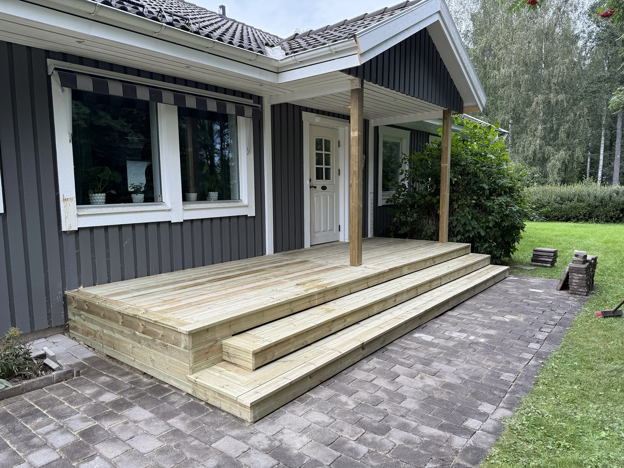 Nybyggt trädäck med trappsteg framför ett grått hus med vitmålade fönster och dörr, omgivet av en grön gräsmatta och träd.