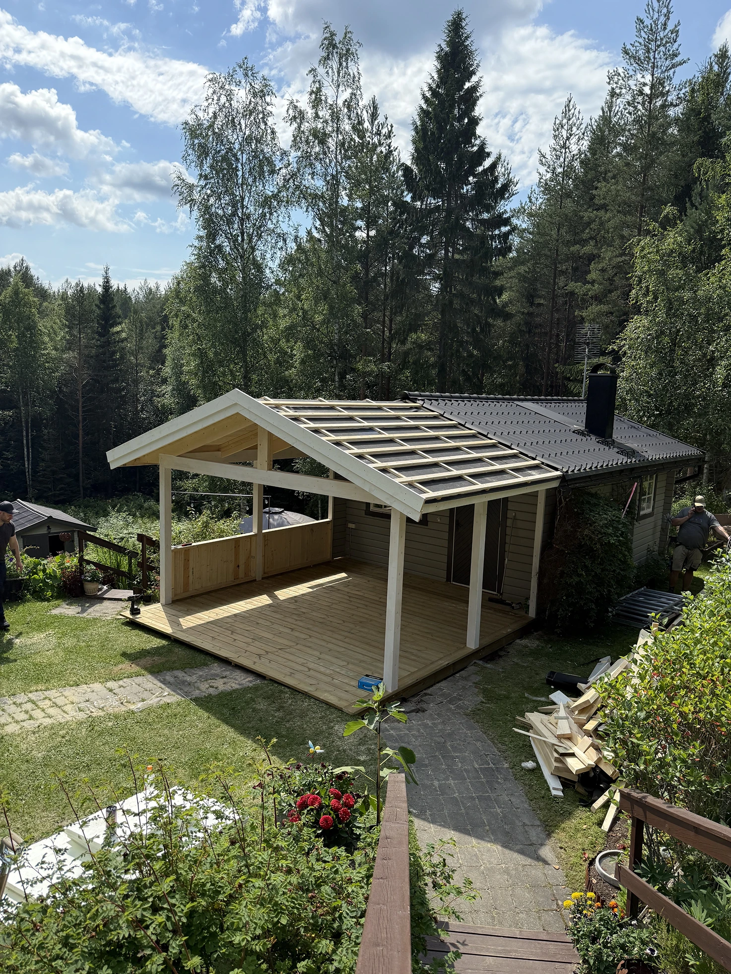 Träterrasskonstruktion under uppbyggnad, fäst vid ett hus, omgiven av gröna träd och trädgårdsblommor.