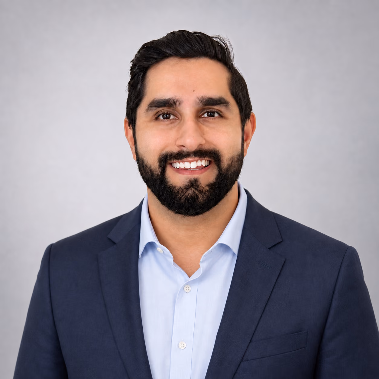 Smiling man with dark hair and beard wearing a navy blazer and light blue shirt against a gray background.