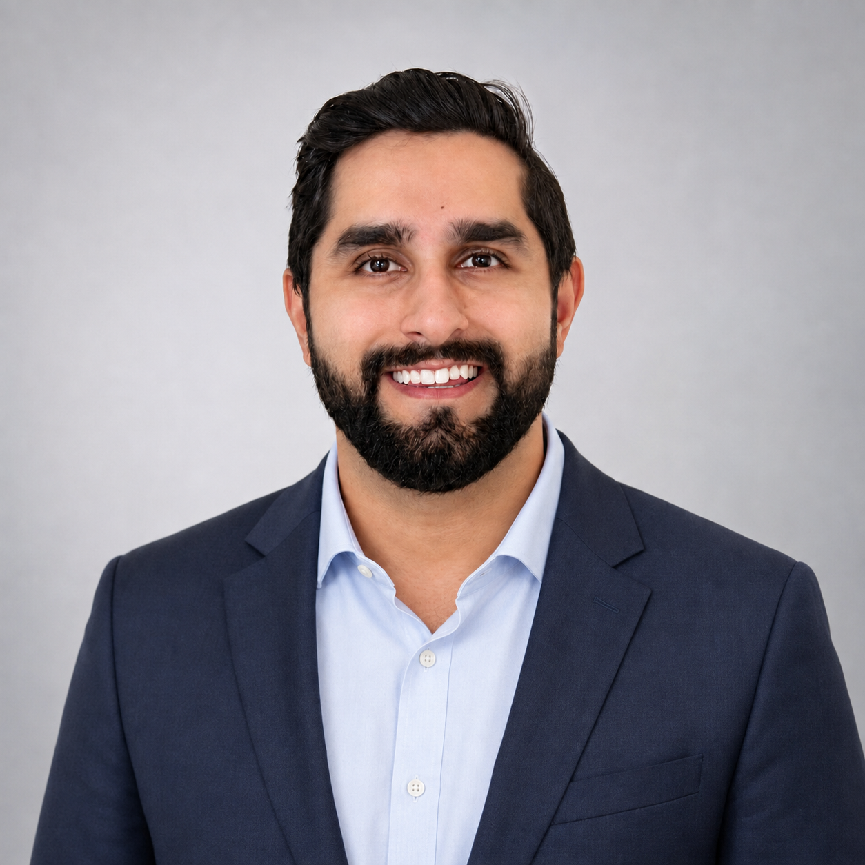 Smiling man with dark hair and beard wearing a navy blazer and light blue shirt against a gray background.