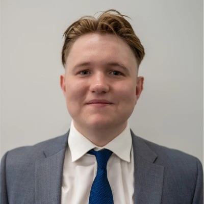Young man wearing a gray suit, white shirt, and blue tie, smiling against a plain background.