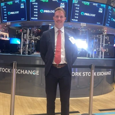 Man in suit and red tie standing at New York Stock Exchange with digital ticker boards behind him.