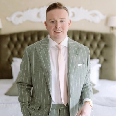 Smiling man in a light gray pinstripe suit with a pale pink tie standing in a bedroom.