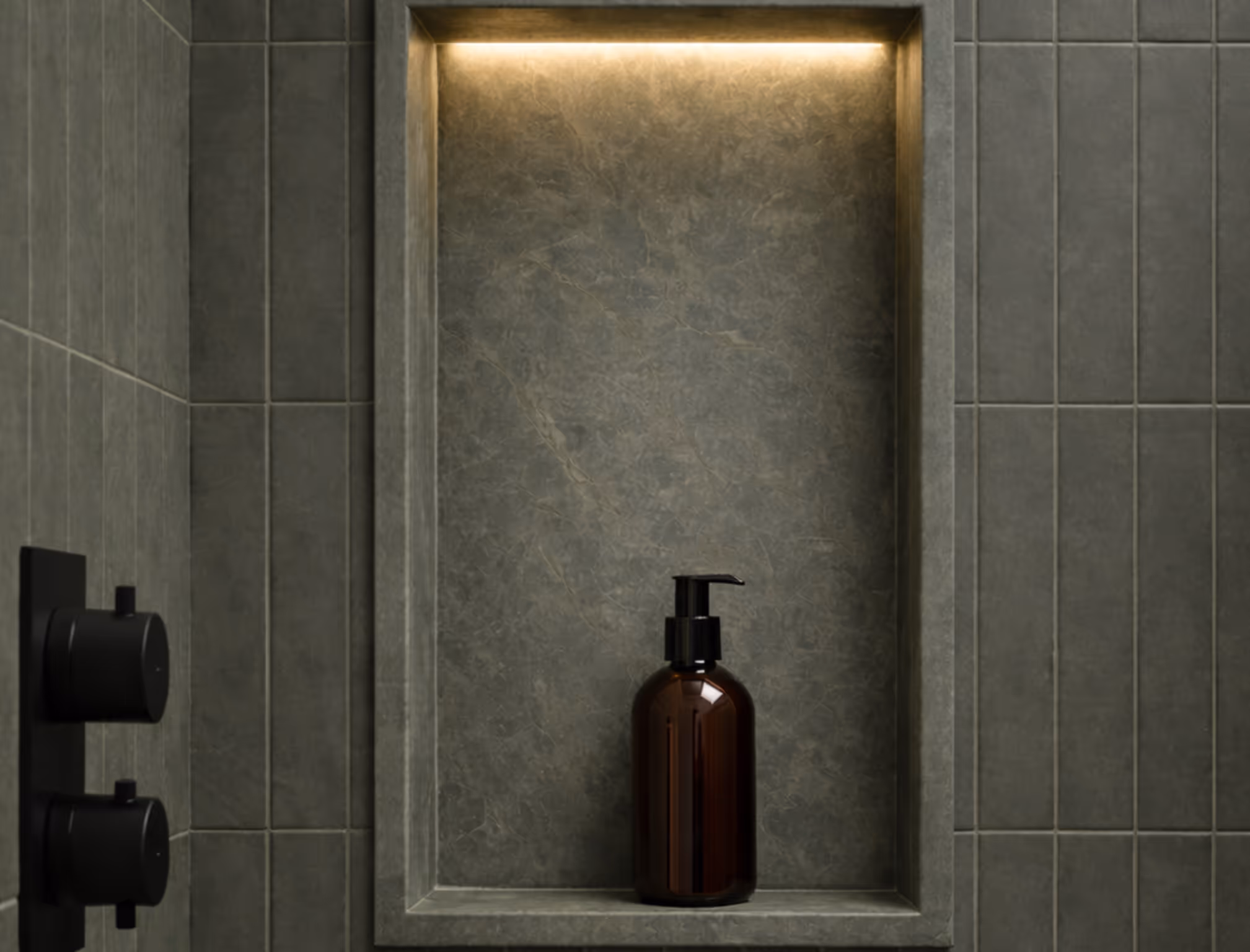 Black pump bottle on a lit recessed shelf in a modern gray tiled bathroom wall.