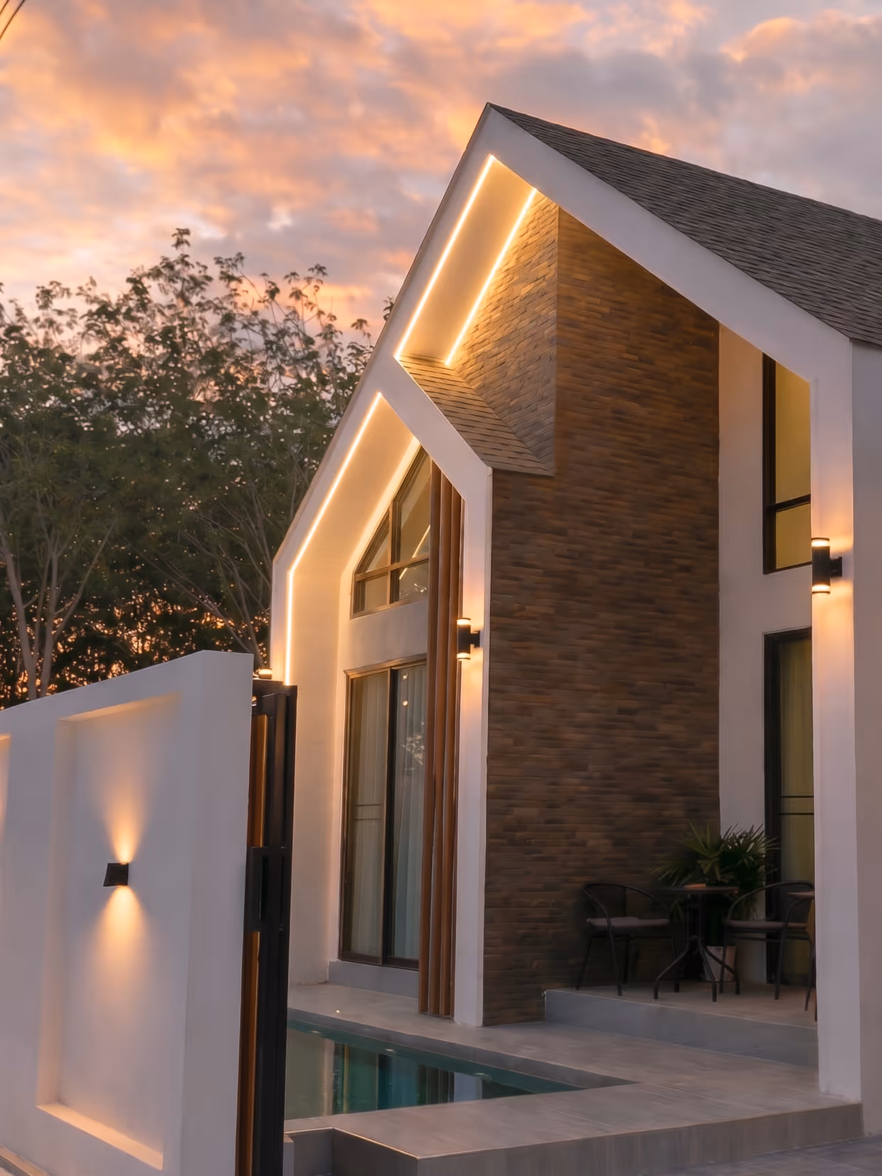 Modern house exterior at sunset with illuminated roofline, large windows, pool, and patio seating.