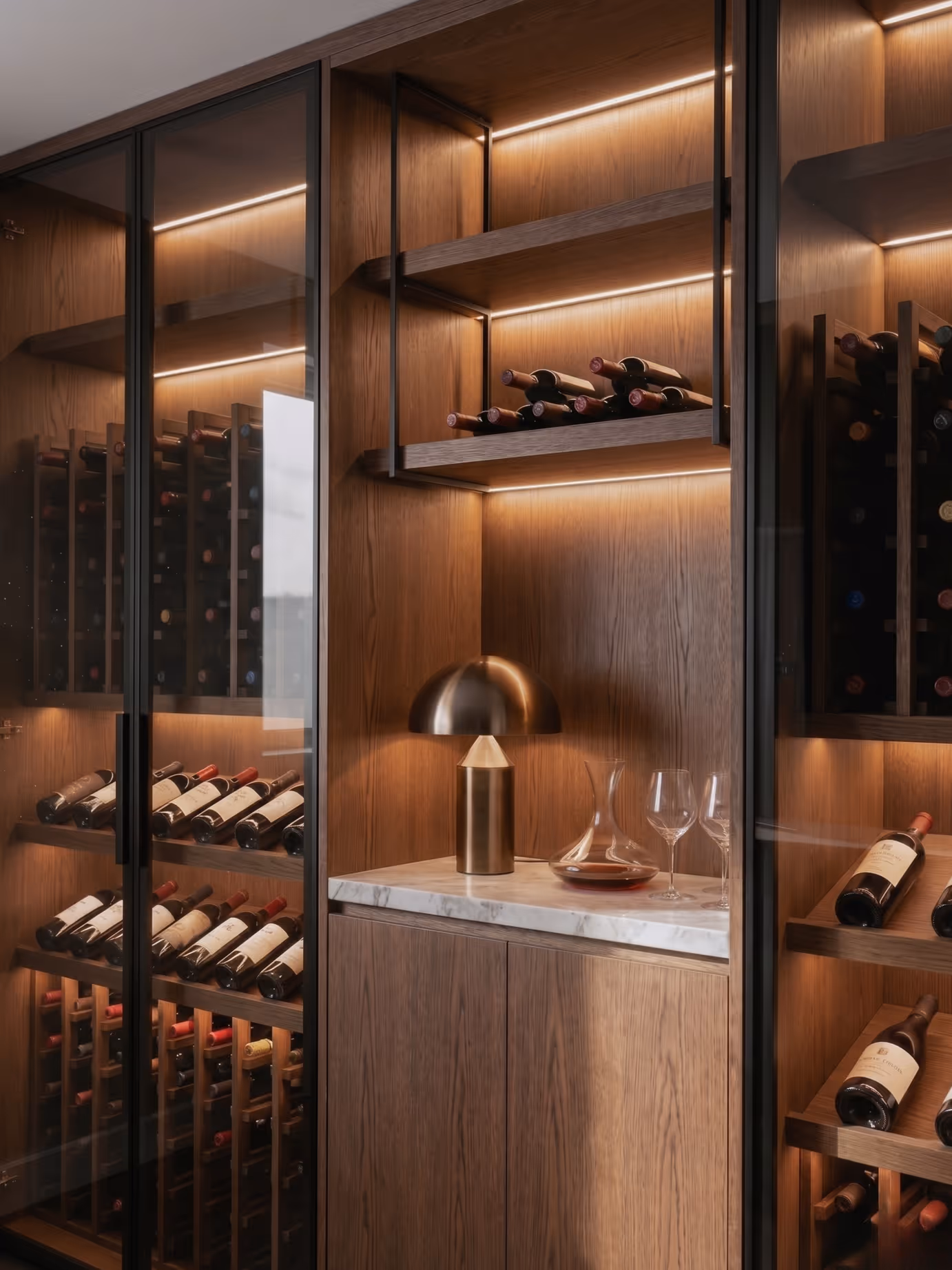 Wooden wine storage with glass doors, bottles, marble counter, lamp, and wine glasses.