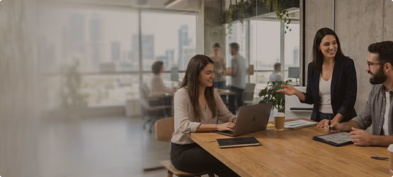 Three coworkers smiling and collaborating around a wooden table with laptops and notebooks in office.