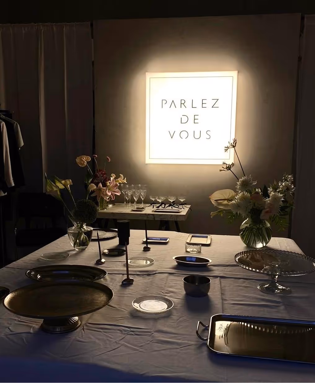 Dimly lit table with plates, flowers, glassware, and a glowing sign reading 'PARLEZ DE VOUS' on the wall.