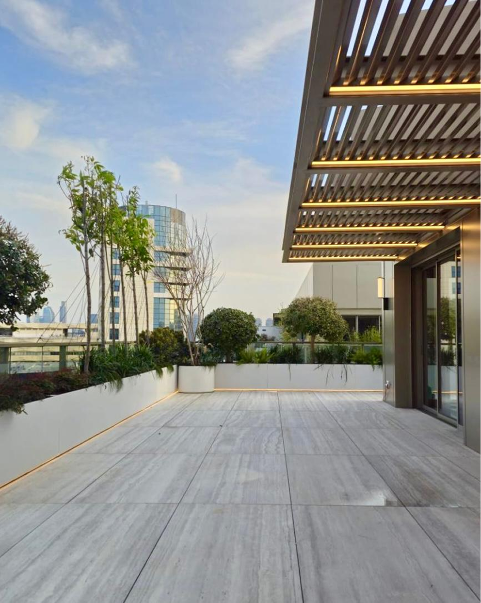 Modern rooftop terrace with large light tiles, plants along edges, and a slatted overhead canopy with lights.