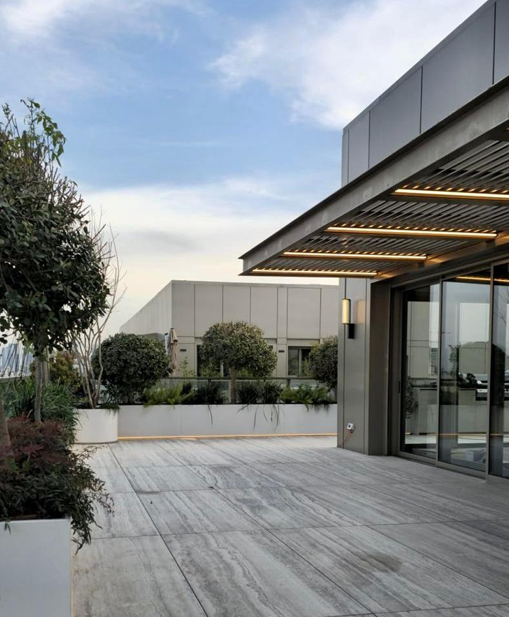 Modern rooftop terrace with potted plants, glass railing, and overhang with embedded lighting.