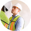 Construction worker wearing a white hard hat and yellow safety vest looking upward.