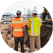 Two construction workers wearing safety vests and helmets discussing plans at a construction site.