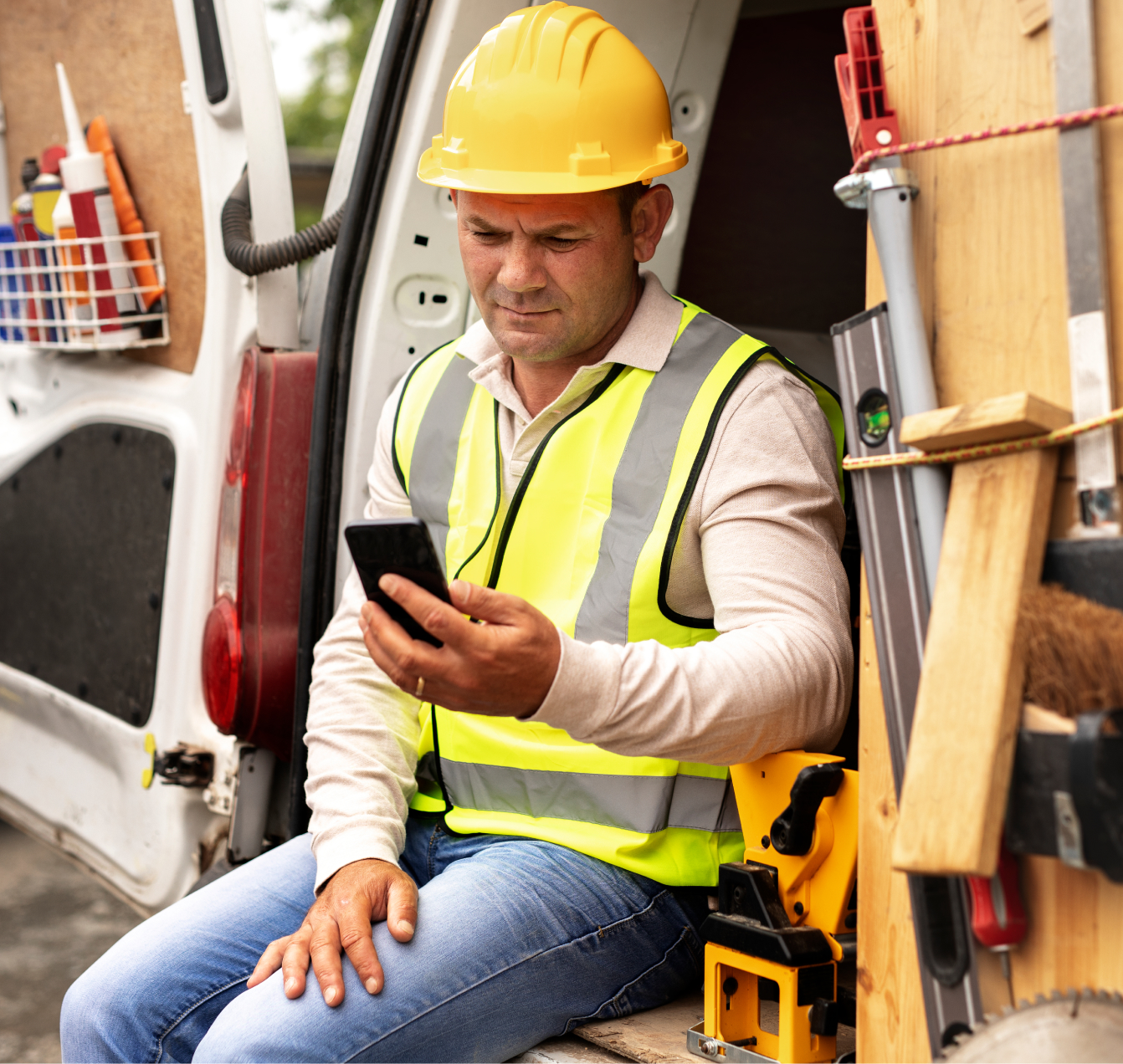 Construction worker wearing a yellow hard hat and safety vest sitting in a van looking at his smartphone.