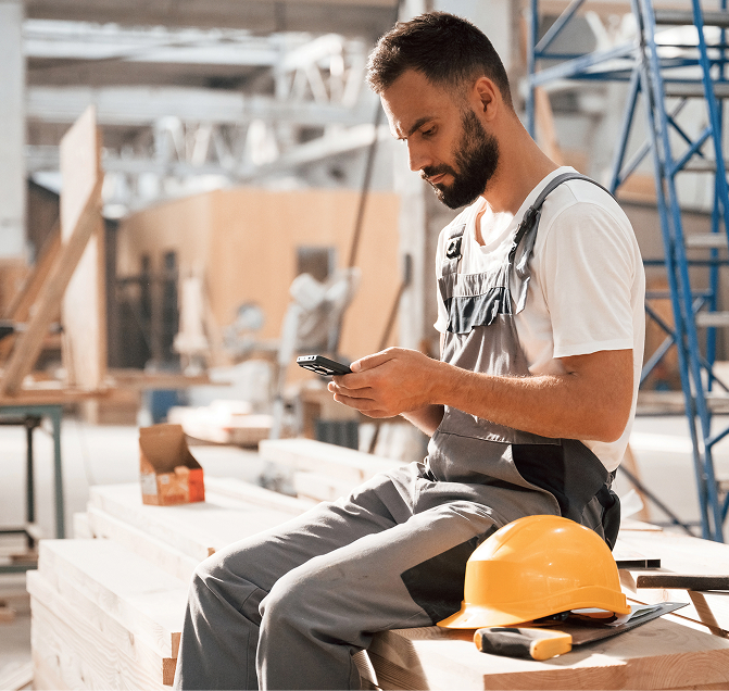 Construction worker in overalls sitting on wooden planks and using a smartphone with a yellow hard hat and tools beside him.