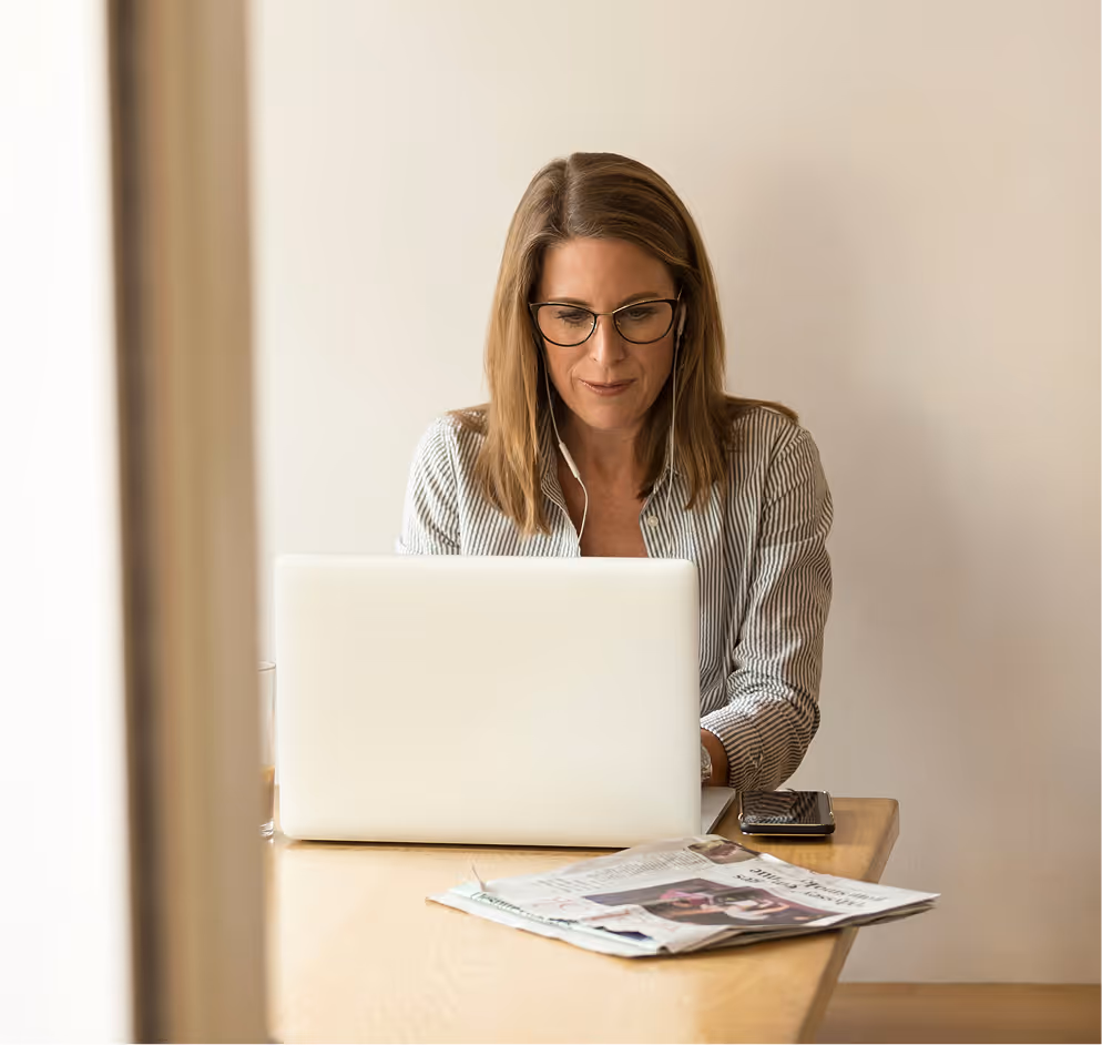 Woman wearing glasses and earphones working on a laptop at a wooden table with a newspaper and smartphone nearby.