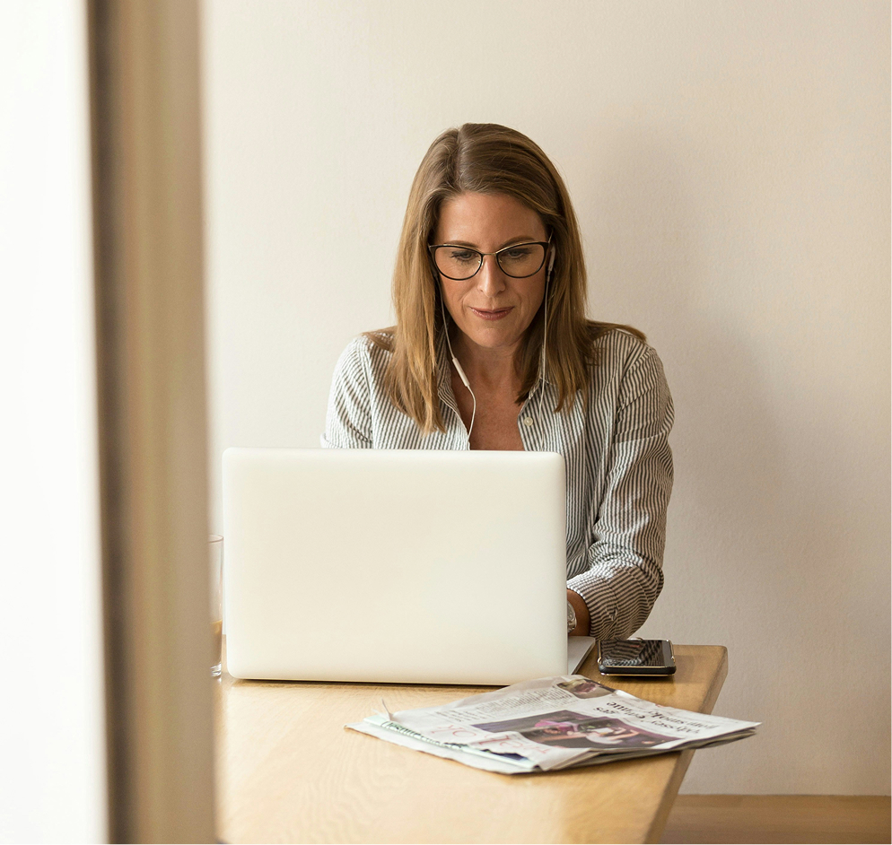 Woman wearing glasses and earphones working on a laptop at a wooden table with a newspaper and smartphone nearby.