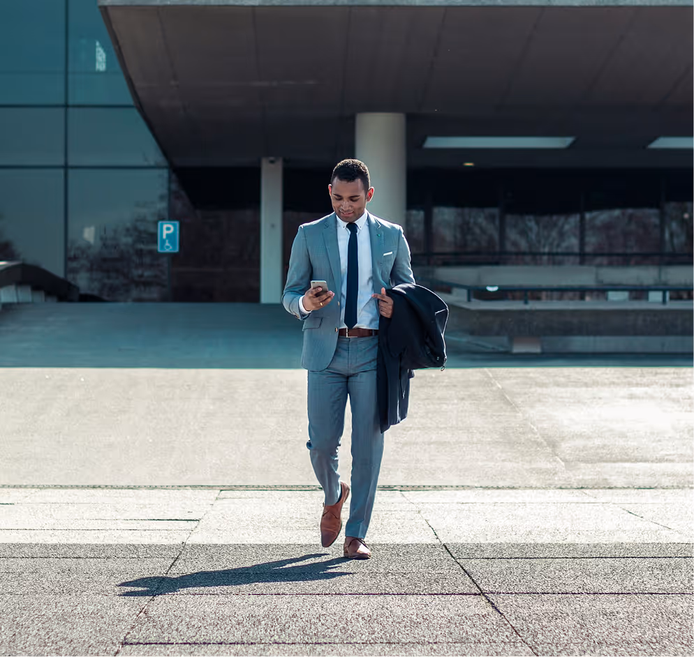 Man in a gray suit walking outdoors while looking at his smartphone and holding a dark jacket.