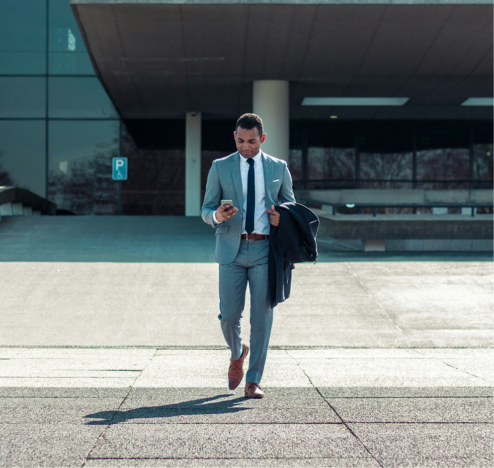 Man in a gray suit walking outdoors while looking at his smartphone and holding a dark jacket.