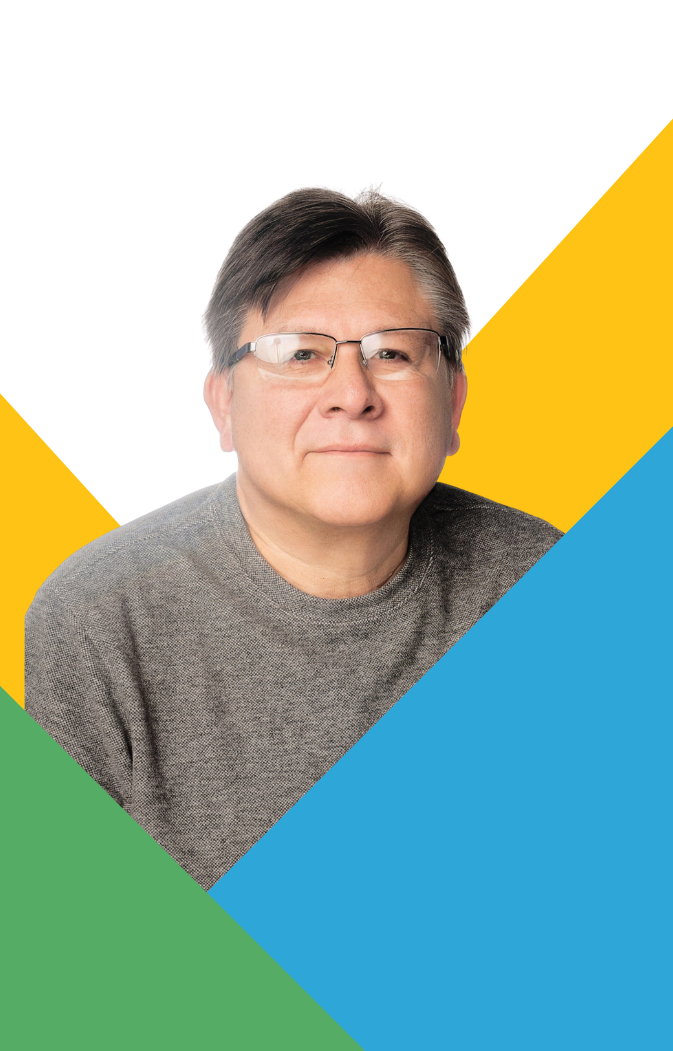 Smiling man with glasses wearing gray sweater, with yellow, blue, and green geometric shapes background.