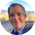 Headshot of a smiling man in a suit with a blurred outdoor background.