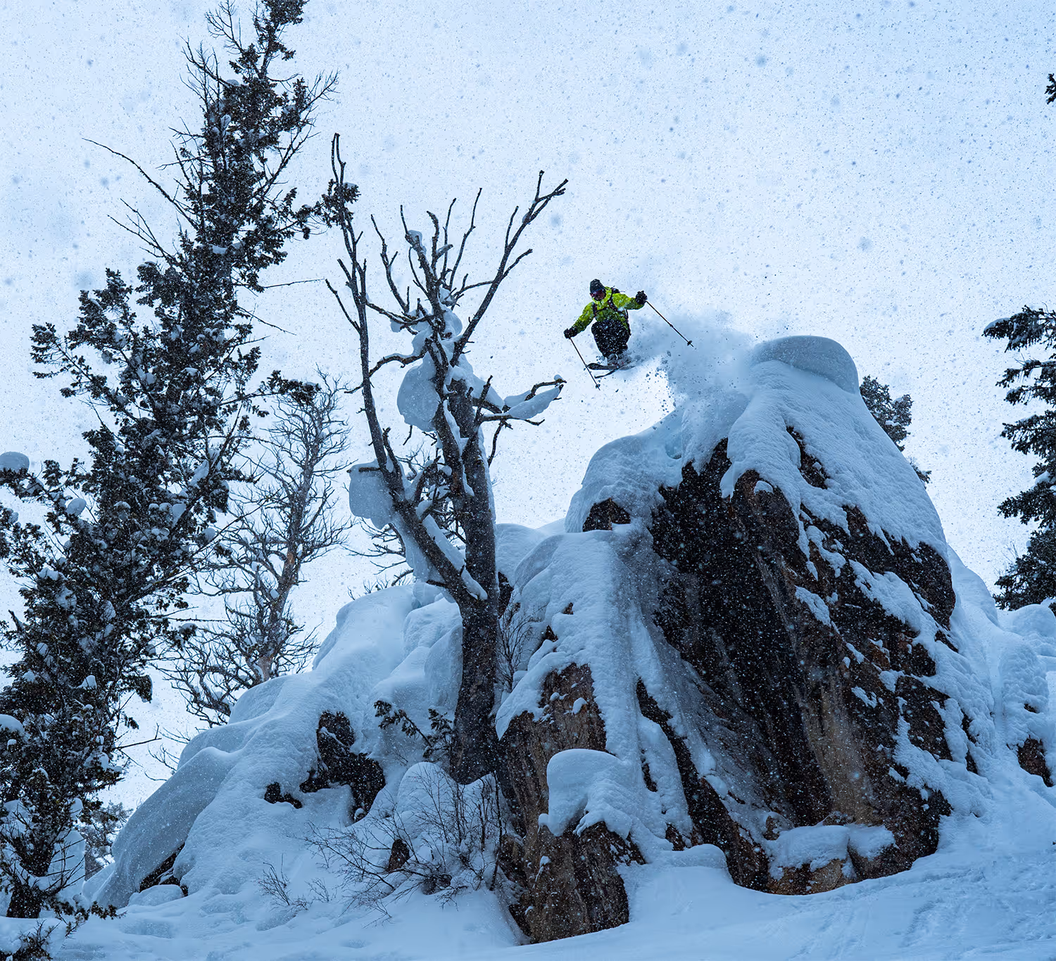 Skier in a bright green jacket airborne above a snow-covered rocky cliff surrounded by snow-laden trees.