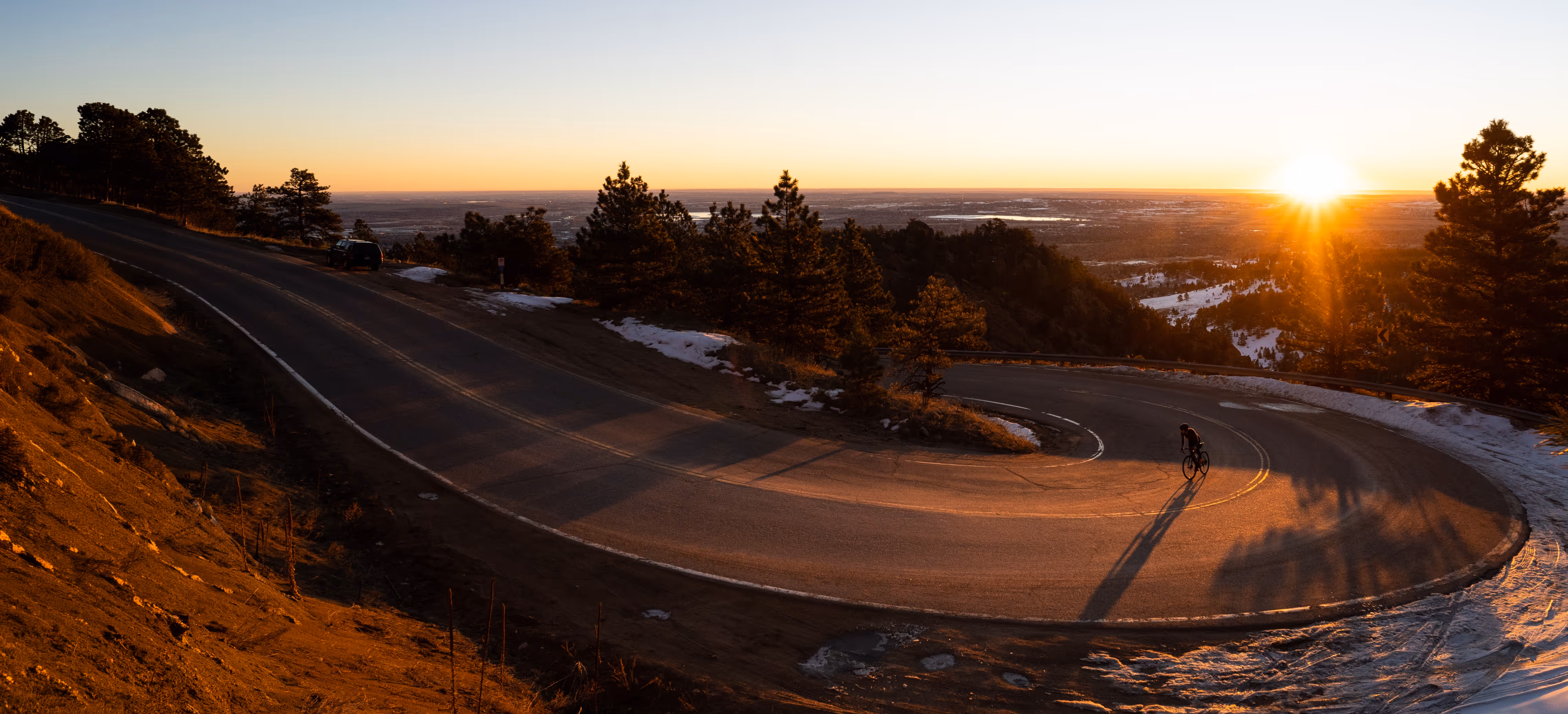 Cyclist riding on a winding mountain road at sunset with trees and patches of snow nearby.