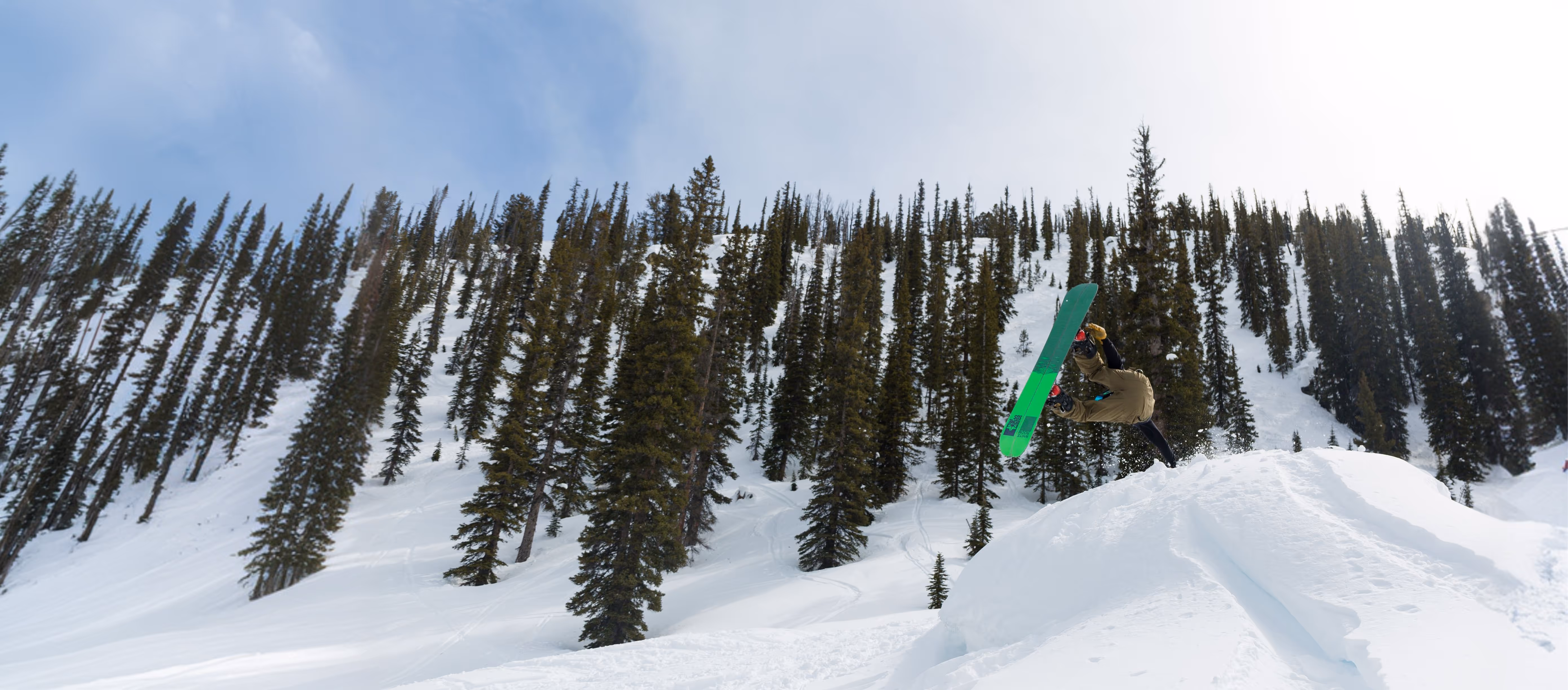 Snowboarder performing a high jump trick on a snowy slope with tall pine trees in the background.