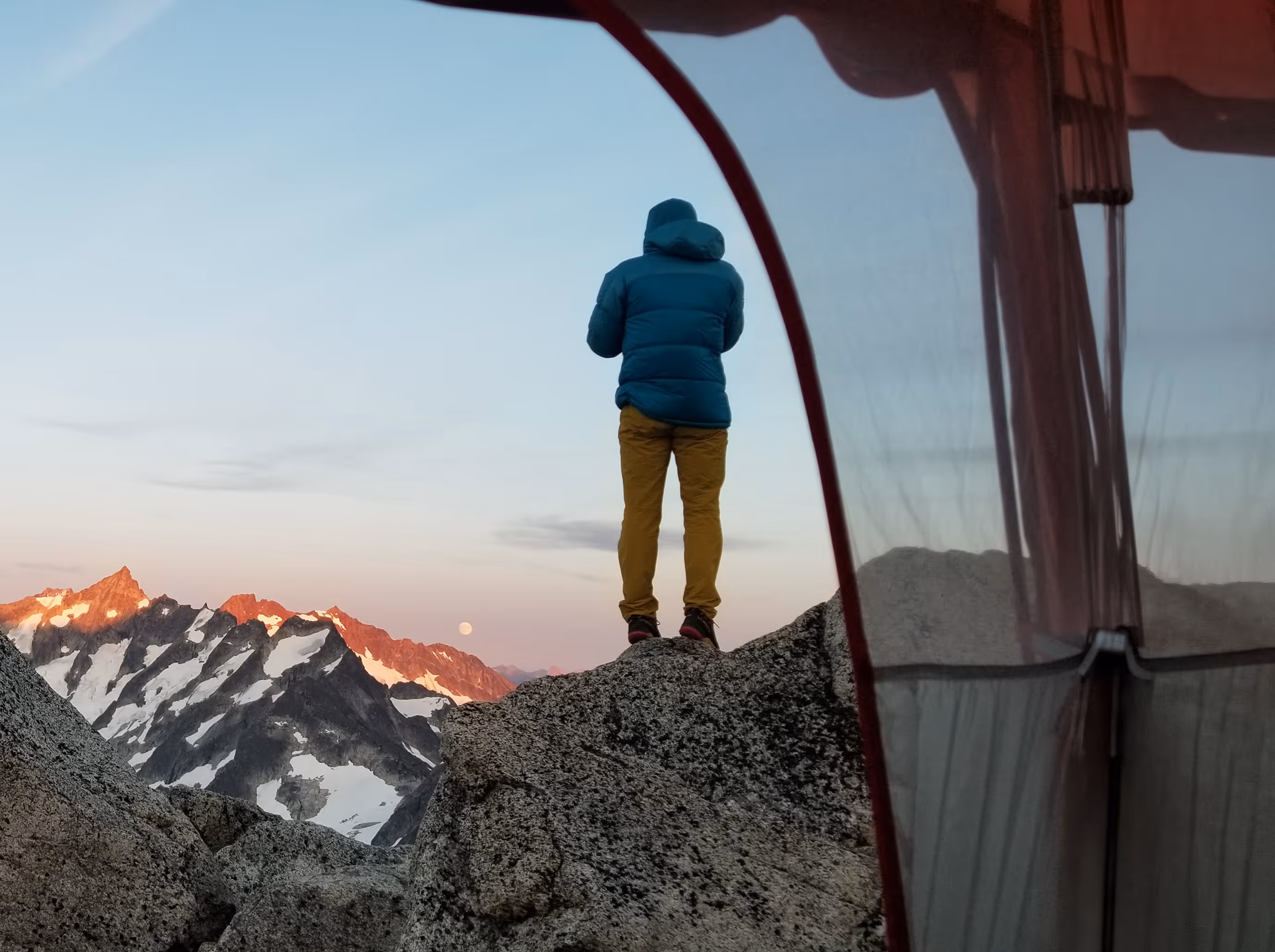 Person in a blue jacket and yellow pants standing on rocky terrain looking at snow-capped mountains at sunset.