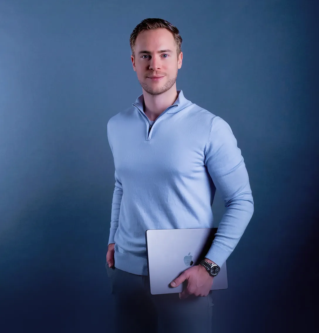 Man in a light blue quarter-zip sweater holding a silver MacBook against a blue background.