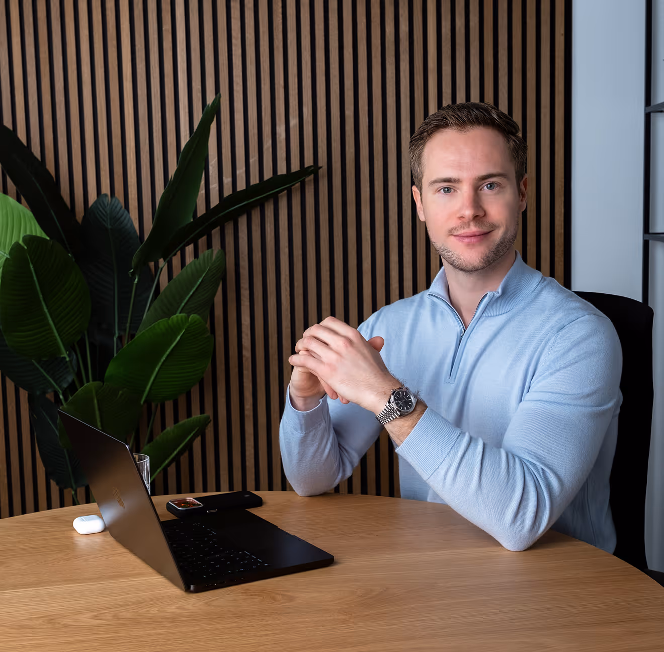 Man in a light blue sweater sitting at a round wooden table with a laptop and wristwatch, smiling at the camera.