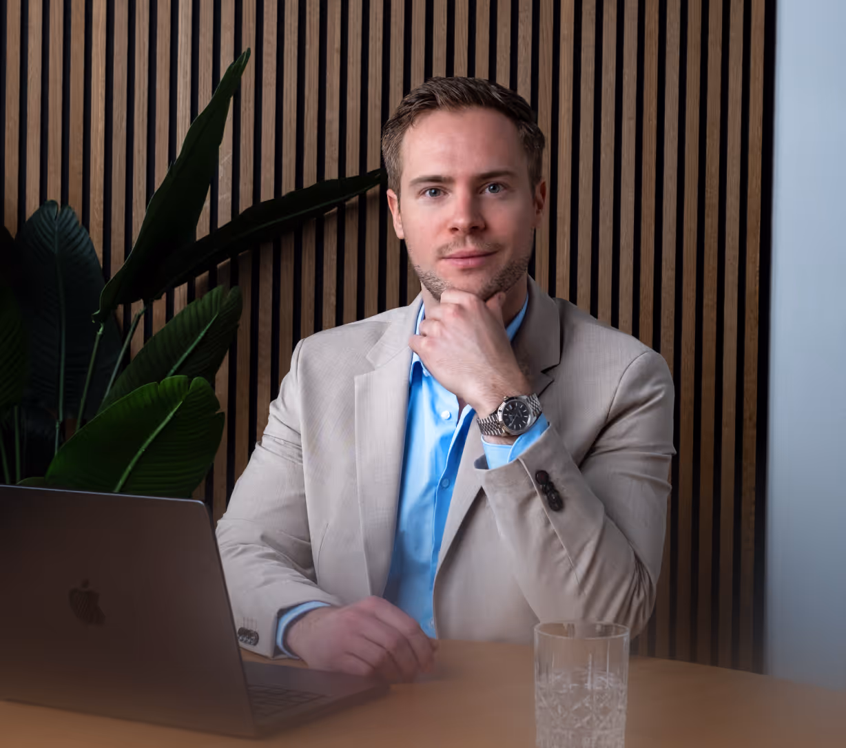 Man in beige blazer and blue shirt sitting at a table with a laptop and glass, resting his chin on his hand.