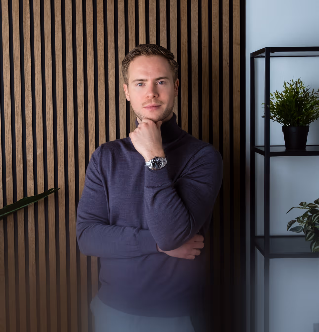 Man in a dark purple turtleneck with a silver wristwatch, standing thoughtfully with hand on chin in front of a vertical wooden panel wall.