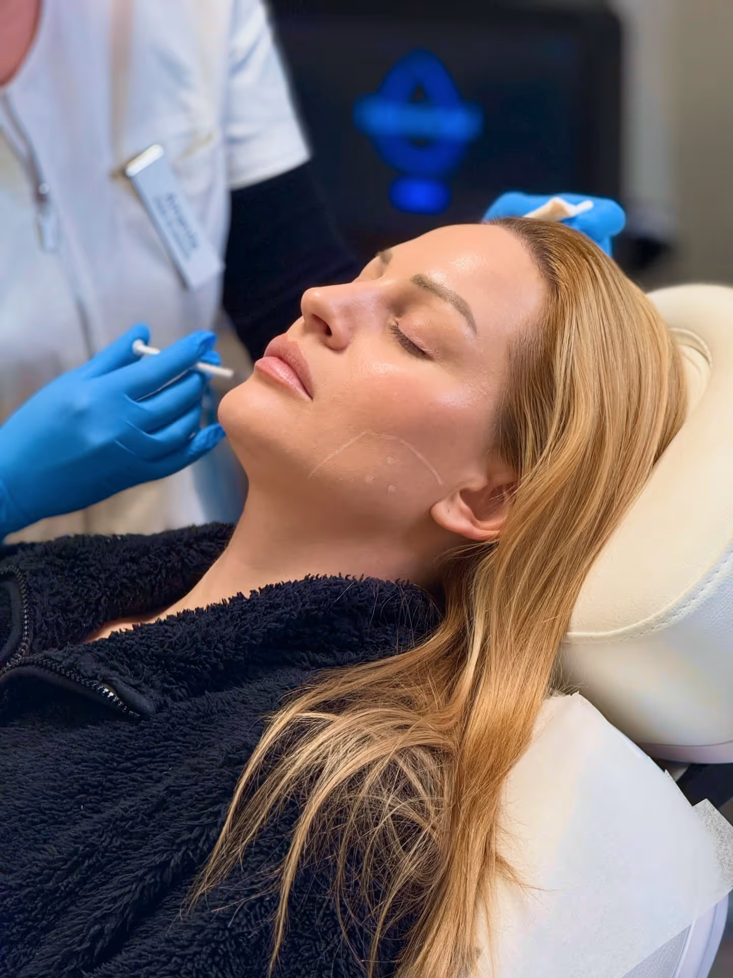 Woman with long blonde hair reclining on a clinic chair receiving facial treatment with blue-gloved hands nearby.