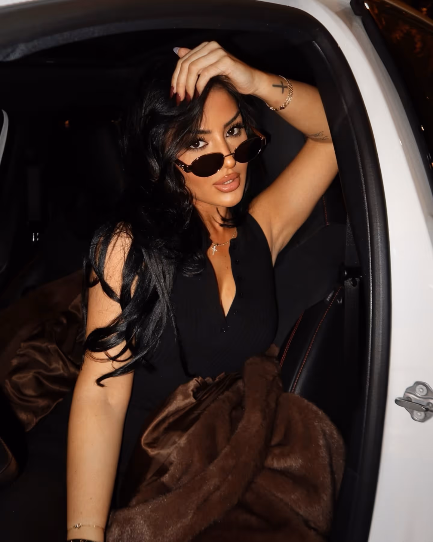 Woman with long black hair and sunglasses poses in a car seat wearing a black top and holding a brown blanket.