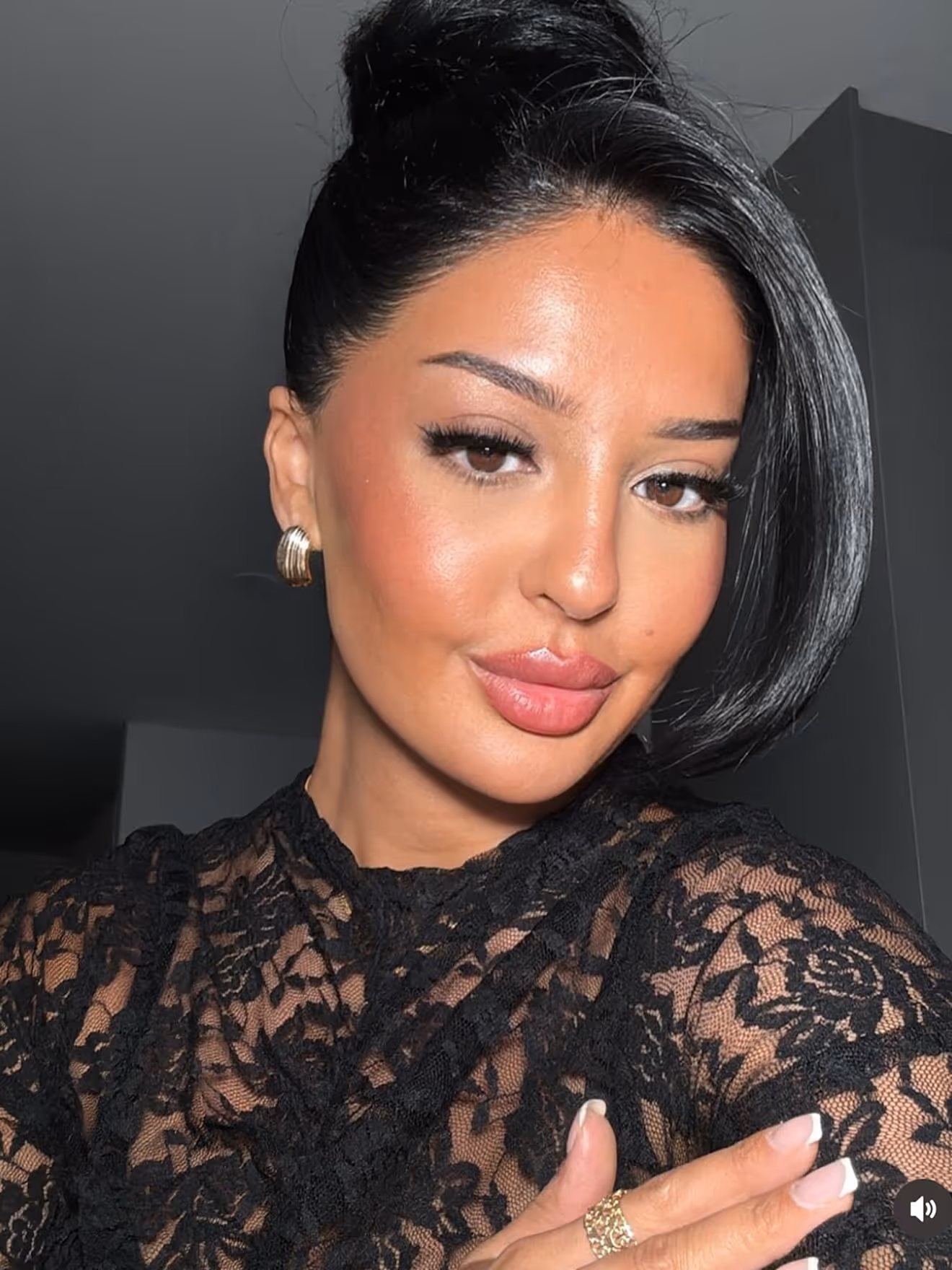 Woman with dark hair styled up wearing a black lace top and gold earrings, posing with crossed arms.