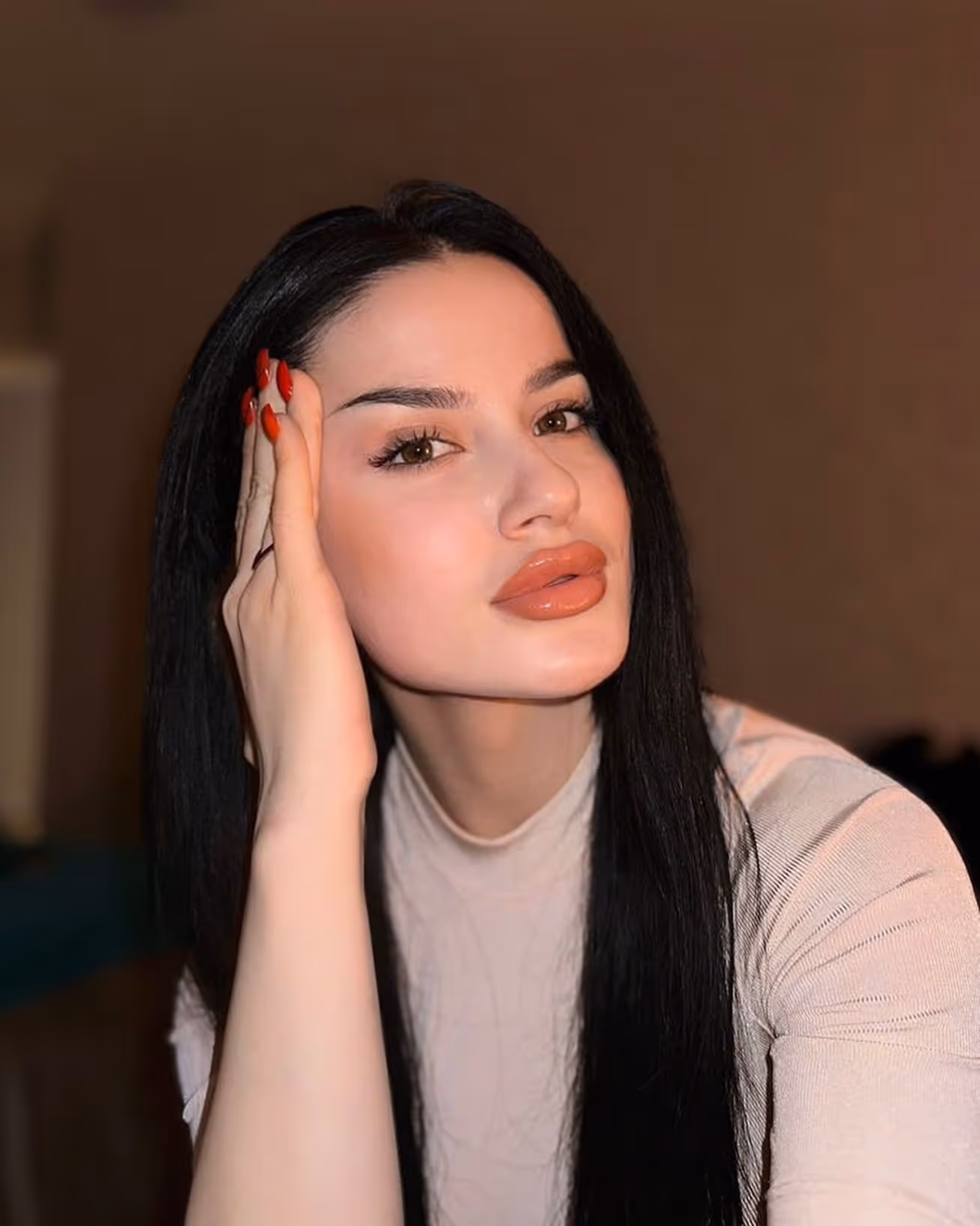 Woman with long black hair resting her face on her hand with red nails and wearing a light beige top.