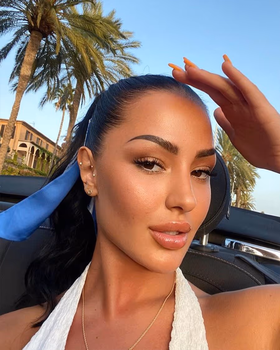 Woman with a white halter top and blue hair scarf posing in a car with palm trees in the background.