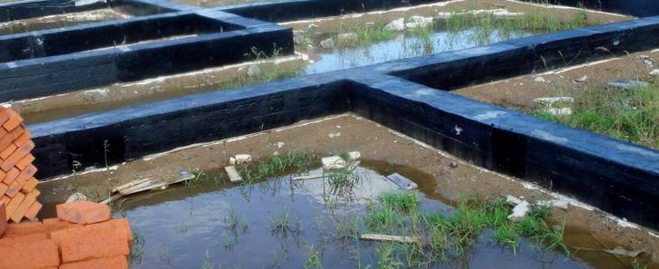 Фундаментні стрічки з темним покриттям на ділянці, між ними стоїть вода; ліворуч видно цеглу