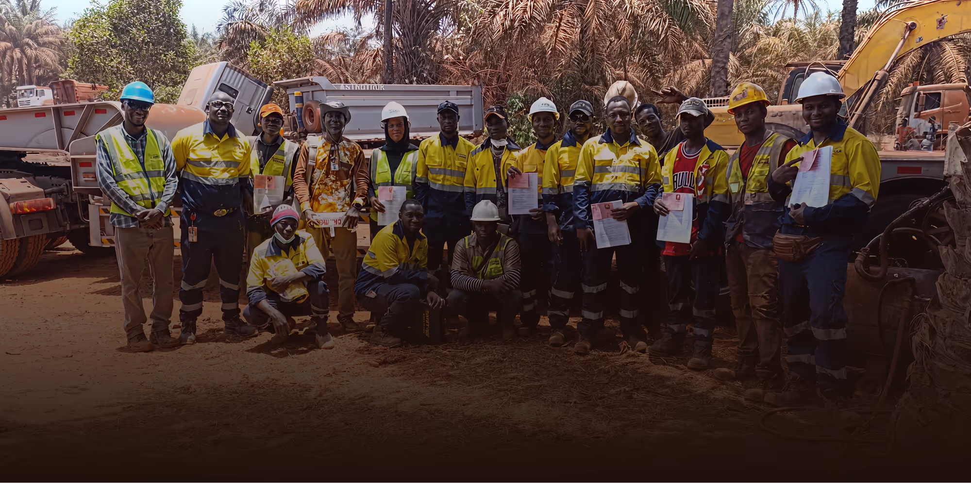 Groupe de travailleurs portant des vêtements de sécurité jaune et casques, posant devant des camions et une excavatrice dans un environnement industriel extérieur.
